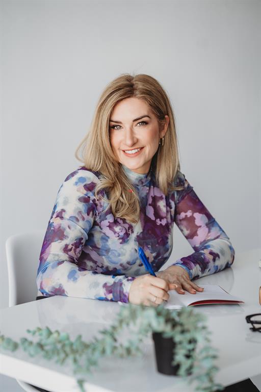 Maude Champagne smiles for a headshot, she is wearing a purple shirt sitting at a desk writing and is  front of a grey background.